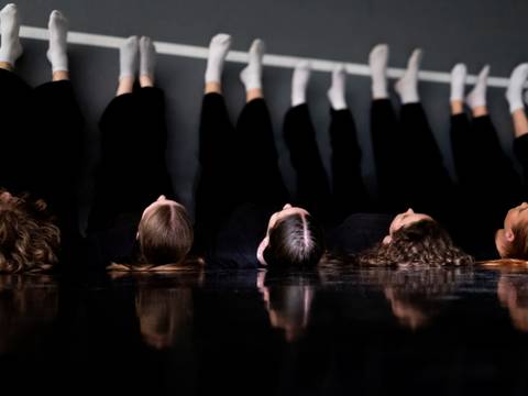 So hart der Fall, so weich die Landung - Ensemble Tanzstudio InRot, Hamburg. – Fünf Personen liegen mit dem Rücken auf dem Boden, der Blick an die Decke gerichtet. Die Beine sind an der Wand angelehnt. Alle tragen graue Socken. An der Wand ist eine weiße Markierung