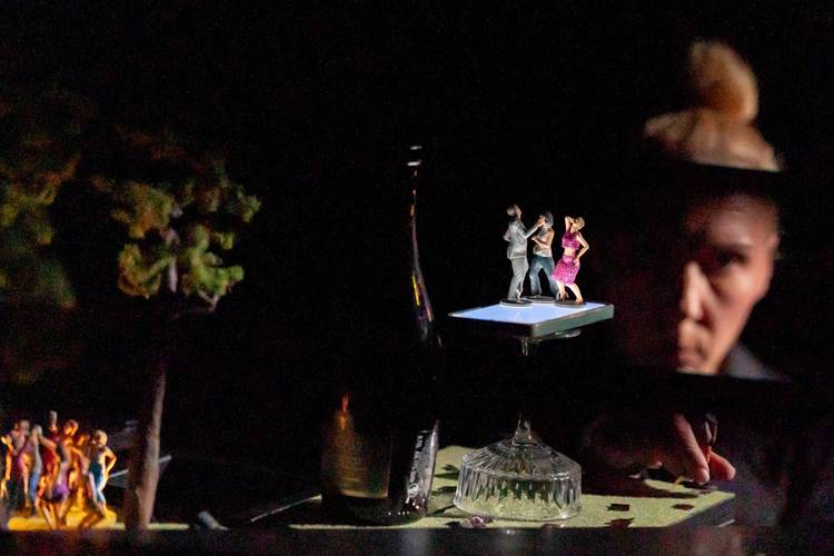 Transport: Fasten your Seatbelts! – Miniaturfiguren tanzen auf einem beleuchteten Podium auf einem Glas. Ein Mensch blickt von rechts, im Hintergrund ist unscharf ein Baum zu erkennen.