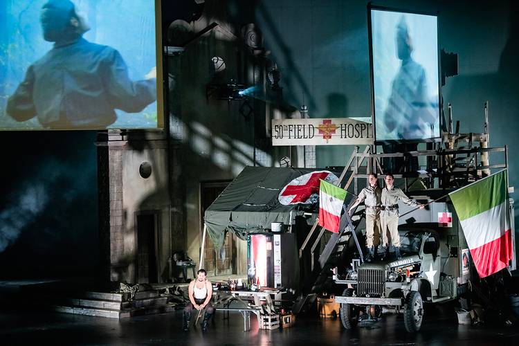 La forza del destino – Zwei Personen stehen auf einem Militärfahrzeug, umgeben von Flaggen. Schild mit "50th Field Hospital", Zelt mit rotem Kreuz, Leinwandbilder im Hintergrund.
