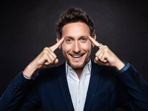 LiorSuchard – Person mit bart lächelt, zeigt mit zeigefingern auf die stirn. Trägt dunkelblauen anzug und weißes hemd. Hintergrund dunkel.