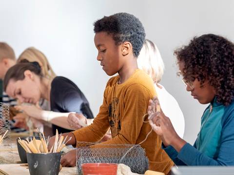  – Gruppe von Personen in einem Workshop arbeitet an Drahtprojekten an einem Tisch. Erwachsene Personen konzentrieren sich. Im Vordergrund ein Kind in gelbem