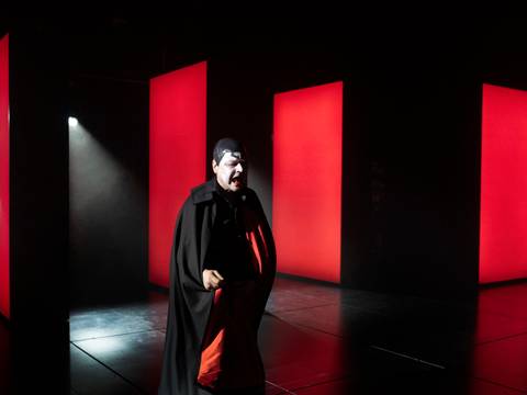 Mephisto – Ein Mann mit weißer Gesichtsschminke und einem rot-schwarzen Umgang steht in einem rot-schwarzem Raum