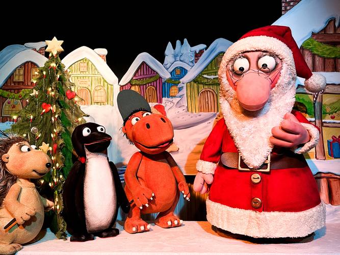 Vier bunte Zeichentrickfiguren vor Weihnachtsbaum und schneebedeckten Häusern; festlich dekoriert mit Herz- und Sternanhängern.