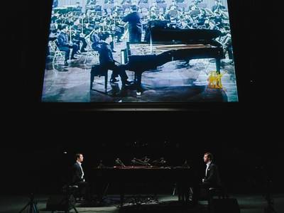 Ming Wong, „Rhapsody in Yellow. Eine Lecture-Performance mit zwei Klavieren“, 2022 – Zwei Pianisten sitzen sich an zwei Klavieren gegenüber. Im Hintergrund befindet sich eine große Leinwand, auf der ebenfalls ein Pianist zu sehen ist, der an einem Flügel sitzt, vor einem Orchester, das ein Dirigent anleitet.