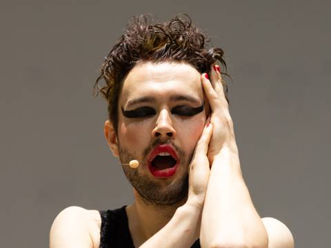 Future is Now – Person mit Make-up, rotem Lippenstift, schwarzem Lidschatten, Mikrofon am Gesicht, trägt ein ärmelloses schwarzes Oberteil, hält Gesicht mit beiden Händen.