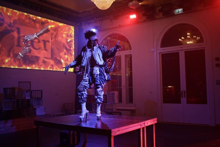 Gira – Portraitperformance Natasha Vergilio – Person in bunter Kleidung und Perücke tanzt auf einem Tisch, im Hintergrund Projektion mit Text "her" und Händen über Flammen. Raum mit roten Lichtern und