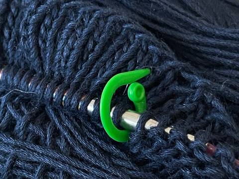 Garn mit Stricknadel und Maschenmarkierer – Zu sehen ist ein dunkelblaues Garnknäuel, in dem eine metallene Stricknadel mit aufgezogenen Maschen steckt. Zwischen zwei Maschen befindet sich ein hellgrüner Maschenmarkierer.