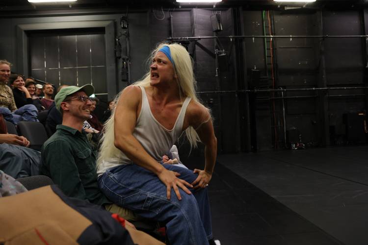 T-Boy on Grindr – Langhaarige Person mit weißem Tanktop und Jeans auf männlich-gelesener Person sitzend, umgeben von lachenden Zuschauer*innen auf Theaterbühne.