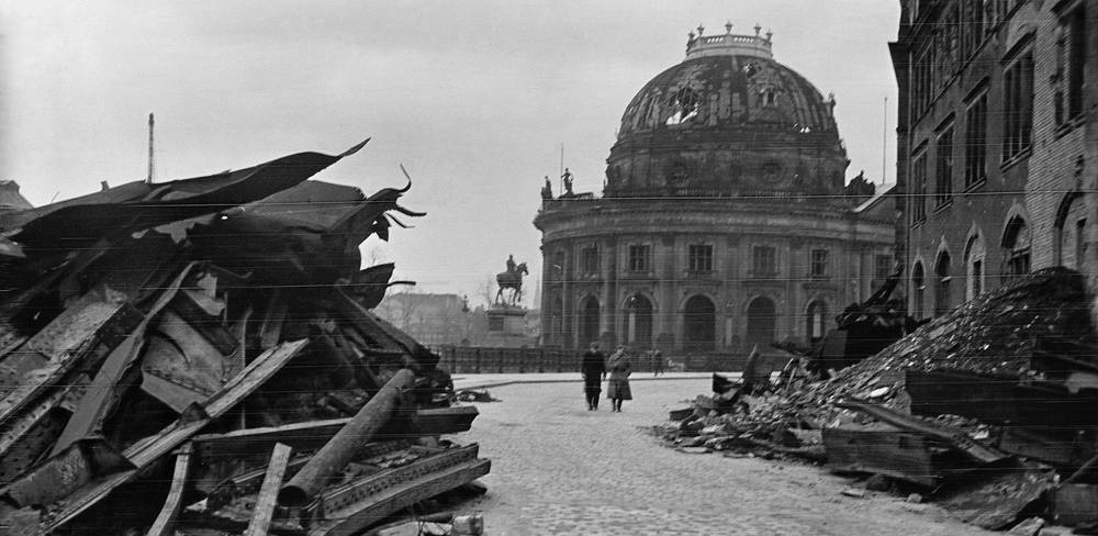 Das zerstörte Kaiser-Friedrich-Museum mit Reiterdenkmal Kaiser Friedrichs III., nach 1945 – Truemmer in einer Strasse; zwei Personen gehen zum Berliner Dom mit eingestuerzter Kuppel; Reiterstatue im Hintergrund; Schwarzweissfotografie.