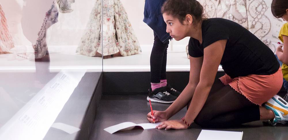 Kind kniet auf Boden mit rotem Stift und Papier vor einer Vitrine mit historischen Kleidern im Museum. Wahrnehmung konzentriert auf Notizen.