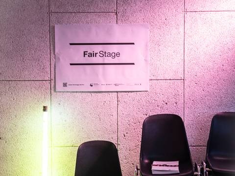  – Gestapelte schwarze Stühle vor hellen Wänden, ein Plakat mit der Aufschrift "FairStage" und eine leuchtende Lampe links.