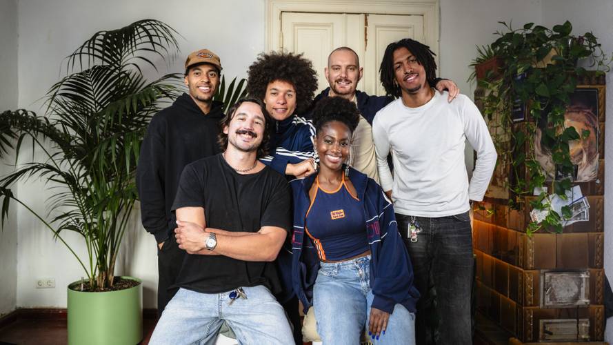 SACODE. Berlin-based collective rooted in afro:diasporic electronic music. – Sechs junge Personen lächeln vor einer Tür. Links steht eine grüne Pflanze.