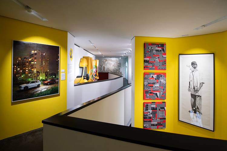 Ein Museumsraum mit gelben Wänden und drei großen Kunstwerken. Eine Fotografie links, geometrische Muster in der Mitte, Zeichnung einer Person rechts.