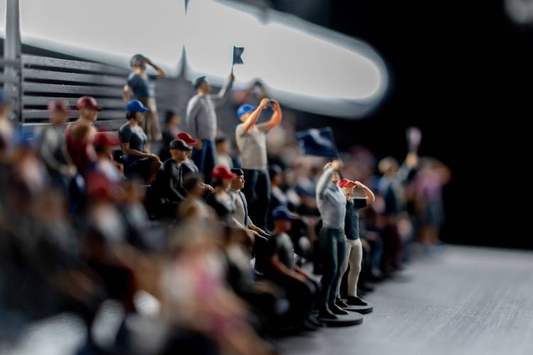Transport: Fasten your Seatbelts! – Verschwommene Figuren in Zuschauertribüne; einige halten Fahnen, tragen Kappen oder machen Fotos. Personen blicken in dieselbe Richtung. Unklare Beleuchtung.