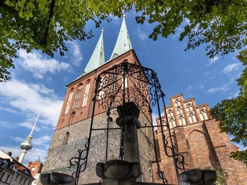 Das Museum Nikolaikirche ist nicht weit von weiteren geschichtsträchtigen Orten Berlins entfernt: dem Rote Rathaus und dem Fernsehturm. – lick durch eine schmiedeeiserne Brunnenzier auf die Türme und Giebel der Nikolaikirche, im Hinterfgrund hinter den Dächern historisierender Gebäude der Fernsehturm und der Turm des Roten Rathauses