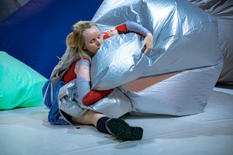 in the air – Person in bunter Kleidung umarmt großes silbernes Kissen, sitzt auf Boden mit buntem Hintergrund aus blauen und grünen Kissen.