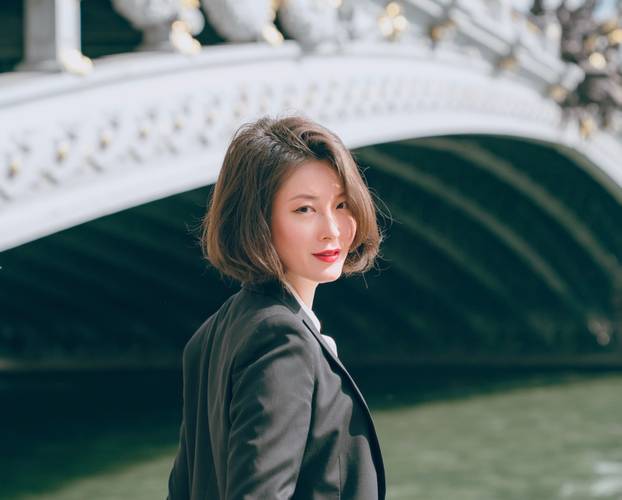 Shin Young Lee – Shin Young Lee mit kurzen braunen Haaren und einem schwarzen Blazer steht leicht lächelnd an einem Fluss, mit einer dekorativen Bogenbrücke im Hintergrund.