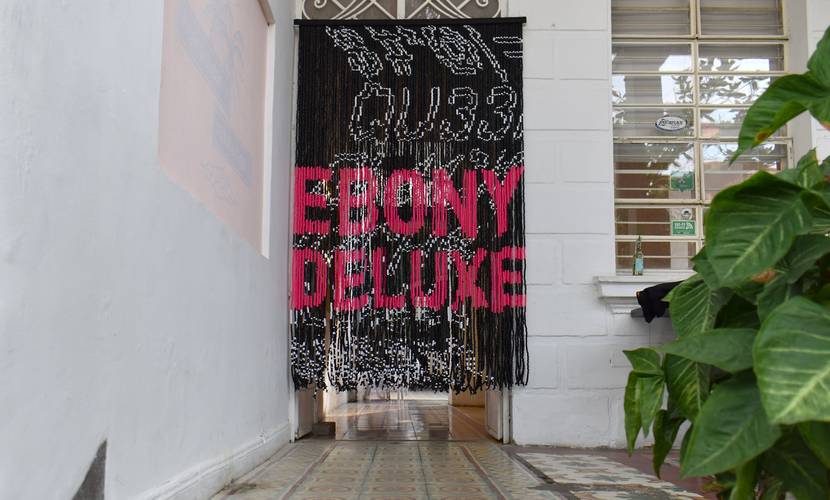Embodied Practices – Talk über Körperwissen 1 – Perlenvorhang mit der Aufschrift „EBONY DELUXE“ in einem Flur mit Pflanzen und gefliestem Boden