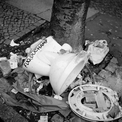 Akinbode Akinbiyi, Neukölln, Berlin, 2019 Aus der Serie: „Photography, Tobacco, Sweets, Condoms, and other Configurations“, seit den 1970er Jahren – Quadratische Schwarz-Weiß-Fotografie: Zu sehen ist eine zerbrochene Toilette und weitere gebrauchte Alltagsgegenstände, die um einen Baum herum verstreut liegen. Auf der Toilette steht in Großbuchstaben mit schwarzem Edding geschrieben „Fuck Duchamp“.