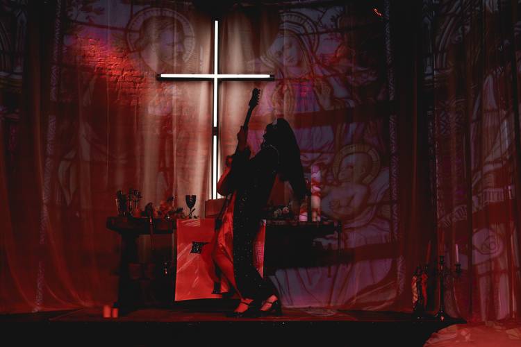 Foto: Michał Wojtarowicz – Person mit Gitarre auf Bühne vor beleuchtetem Kreuz, umgeben von religiösen Symbolen. Tisch mit Kerzen und Schalen links. Roter Lichtschein.