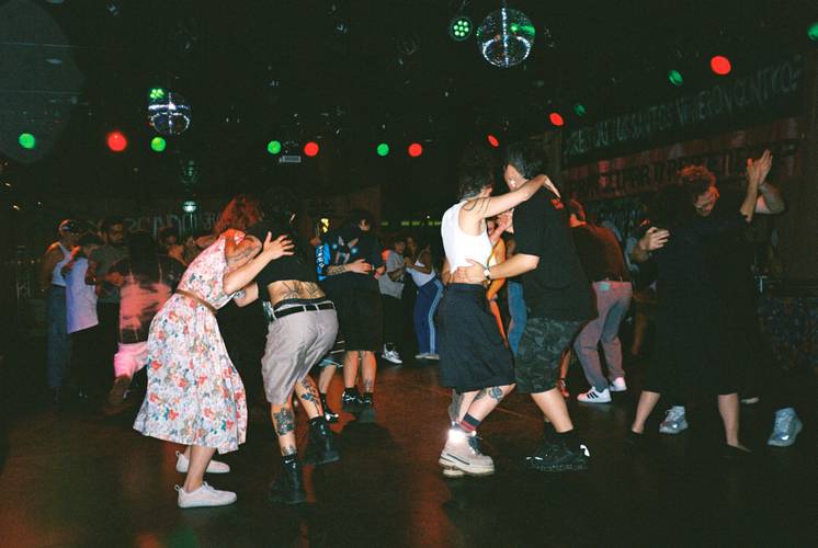 Menschen tanzen eng umschlungen in einem dunklen Raum mit Discokugeln und bunten Lichtern. Viele tragen dunkle Kleidung und haben Tattoos.