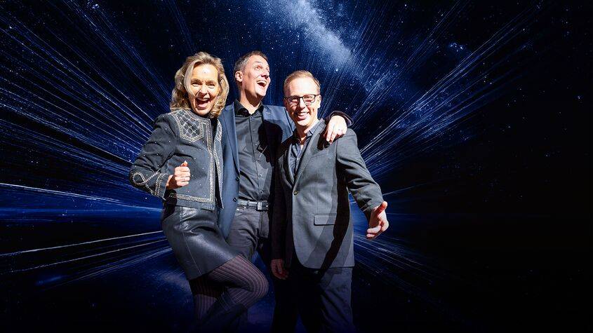 Drei gut gelaunte Personen in eleganter Kleidung vor einem Hintergrund mit Weltraum-Motiv. Alle lachen und machen Daumen-hoch-Gestik.