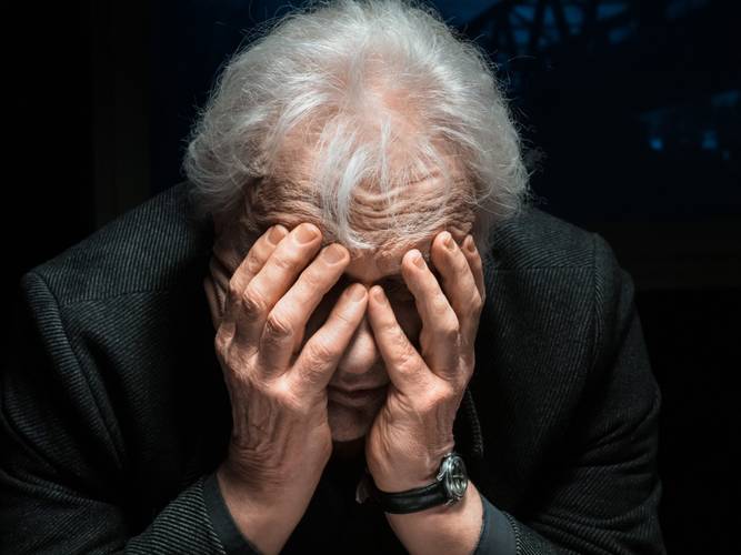 Eine ältere Person mit grauen Haaren hält den Kopf in die Hände, trägt einen dunklen Anzug und eine Uhr am linken Handgelenk.