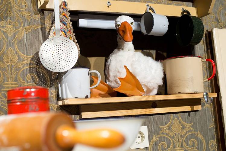 Kuscheltier-Gans auf einem Regal zwischen Emaillebechern, daneben ein Sieb und ein Nudelholz vor einer dekorativen Tapete.