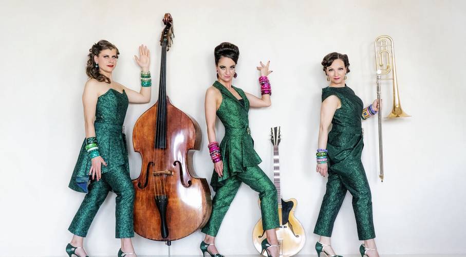 Zucchini Sistaz „Eleganz oder gar nicht!“ – Drei Personen in grünen Anzügen, von links nach rechts: Kontrabass, Gitarre und Posaune. Armbänder in verschiedenen Farben, High Heels.
