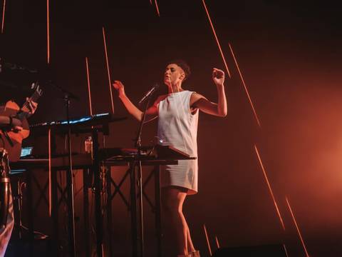  – Zwei Personen auf der Bühne, eine spielt Gitarre, die andere singt am Mikrofon. Hintergrund mit orangenen Lichtstrahlen.