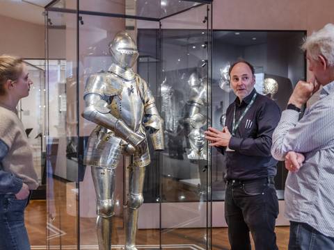  – Ein Museumsführer steht vor einer Ritterrüstung in einer Vitrine und erklärt sie zwei Personen.
