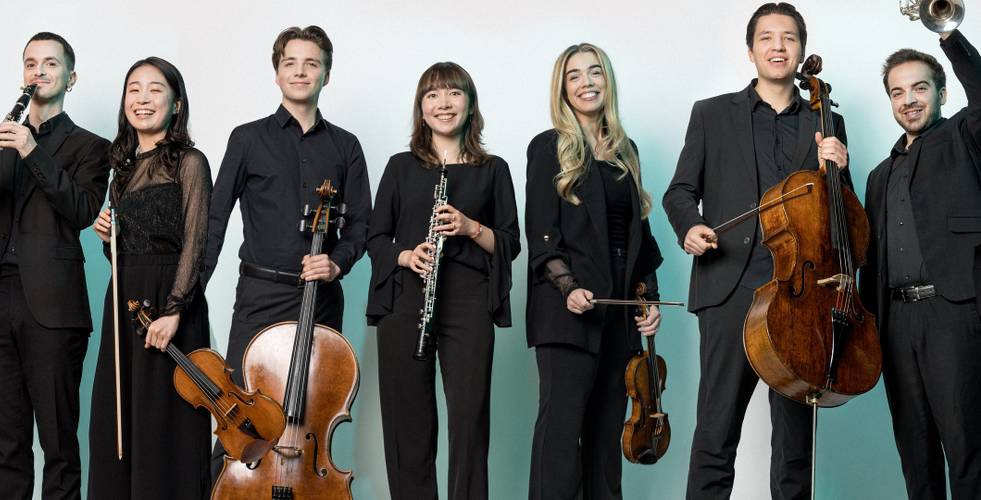 Sieben Personen mit Musikinstrumenten stehen nebeneinander: Klarinette, Violine, Violoncello, Oboe, Violine, Violoncello und Trompete. Alle tragen schwarze