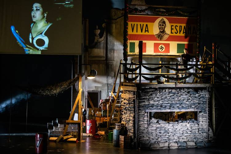 La forza del destino – Große Bühne mit Projektion einer Frau. Schild mit „Viva Espana“, Bild eines Soldaten, Flaggensymbole mit Hakenkreuz. Person auf Gerüst.