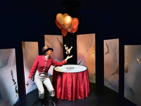 – Person in roter Jacke und Hut hält Luftballons. Tisch mit rotem Tuch, umgeben von bemalten Leinwänden.