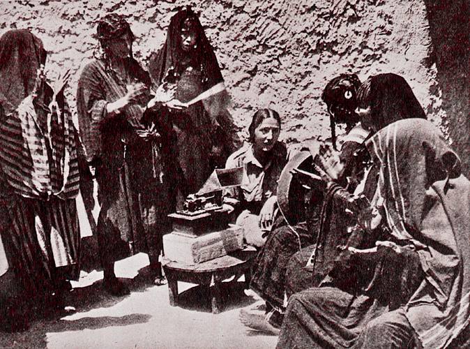 Foto aus Brigitte Schiffers Dissertation "Die Oase Siwa und ihre Musik" (1936) – Sechs verschleierte Personen stehen und sitzen um eine Kamera auf einem Tisch vor einer Steinwand.