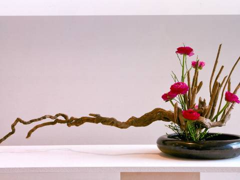 Ikebana – die Kunst des Blumensteckens – Ikebana-Arrangement mit roten Blumen und Ästen in schwarzer, runder Vase auf weißem Tisch vor hellem Hintergrund.